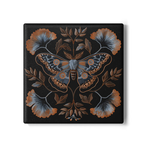 Square black ceramic coaster with detailed copper and blue moth illustration centered among symmetrical botanical elements including ginkgo-like leaves and delicate foliage.
